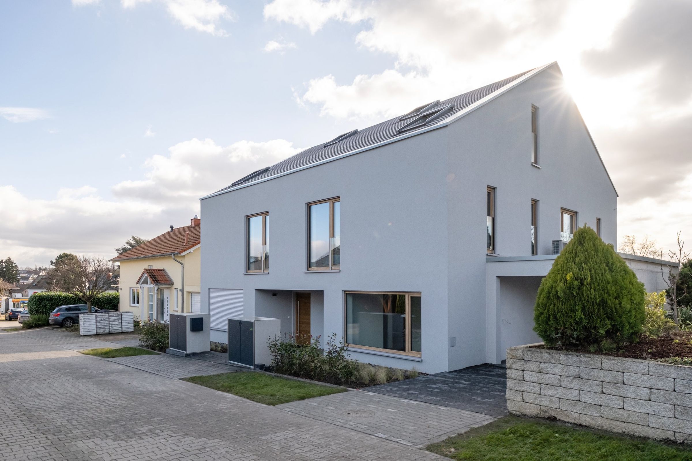 Haus M in Mainz: helle Putzfassade mit Satteldach im Gegenlicht, davor Pflaster und Trockenmauer