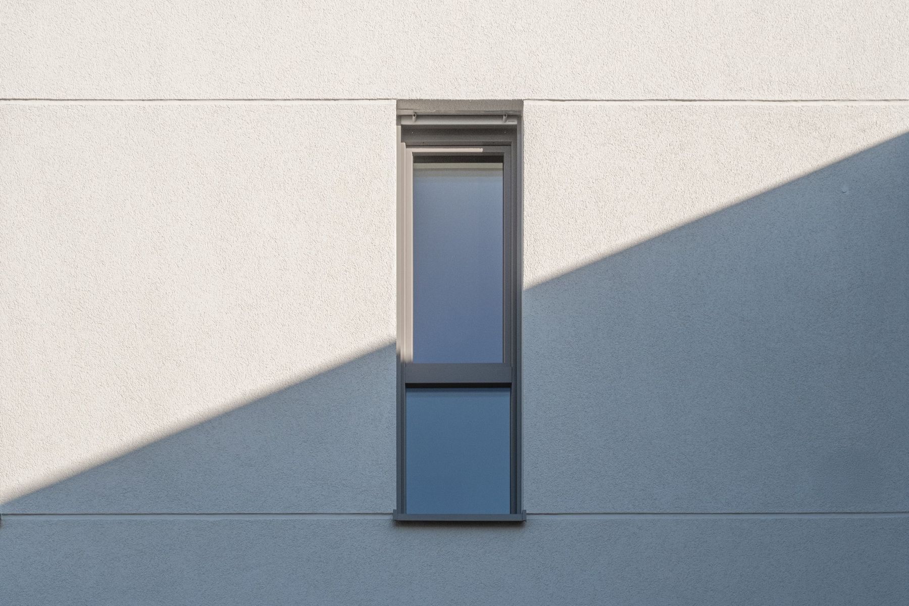 Mehrfamilienhaus K, schmales Fenster in heller Putzfassade mit Schattenkante